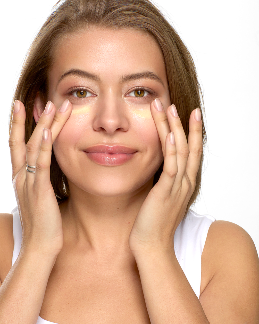 A bright, front-facing portrait of a smiling woman with her hair pulled back, using her ring fingers to gently pat cream into the skin under her eyes, demonstrating a delicate skincare application.