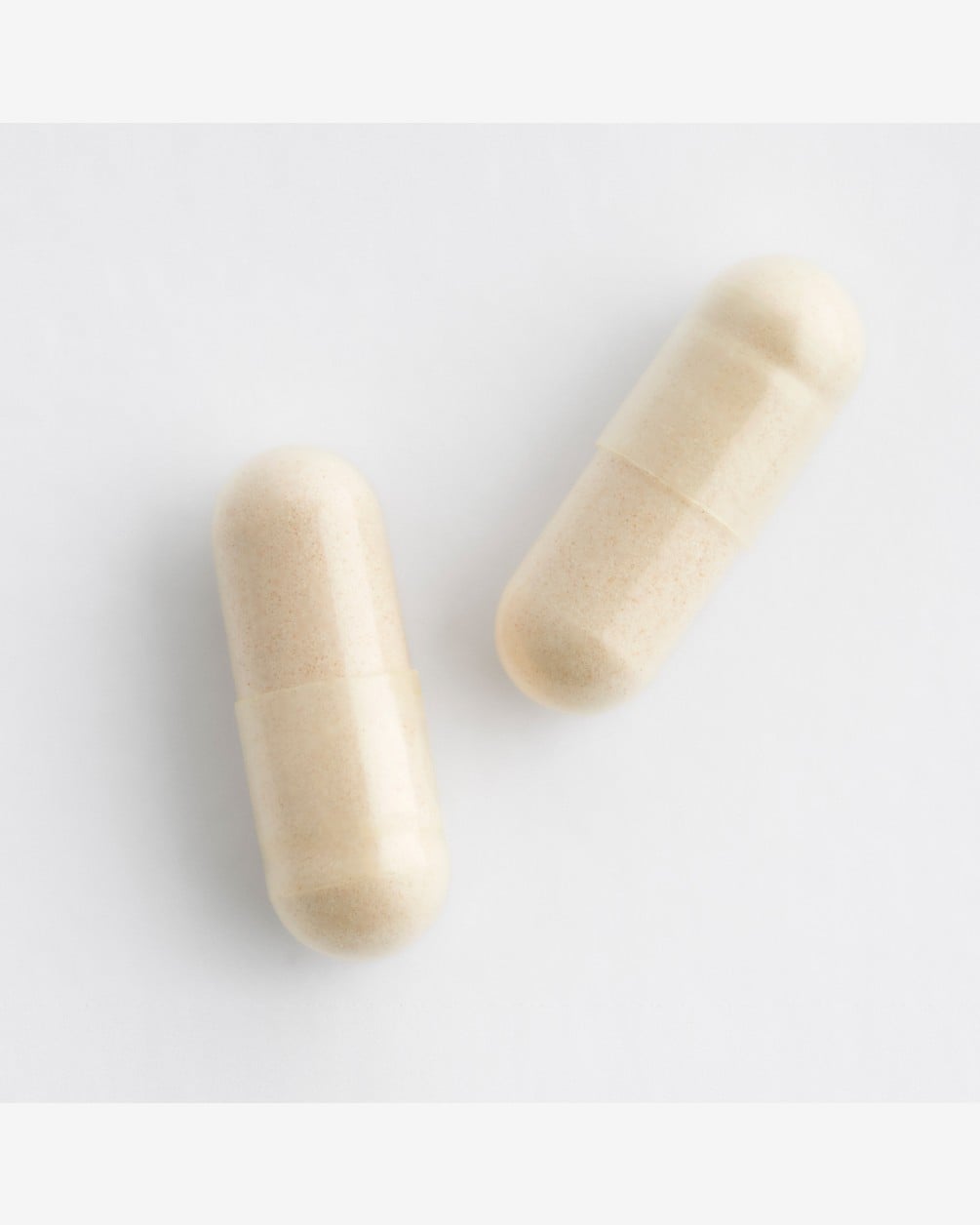 A close-up, top-down shot of two white supplement capsules resting on a plain white surface. The capsules have a standard oblong shape and a slightly textured appearance.