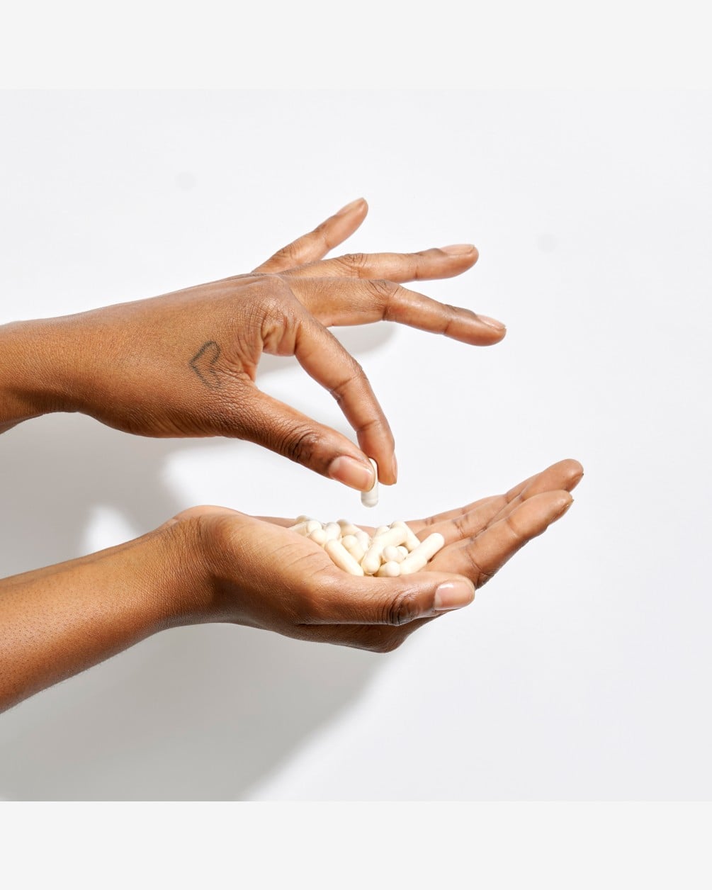 A close-up shot of two hands against a white background. One hand is held open, filled with several white capsules, while the other hand is shown mid-motion, picking up or dropping a single capsule into the palm.
