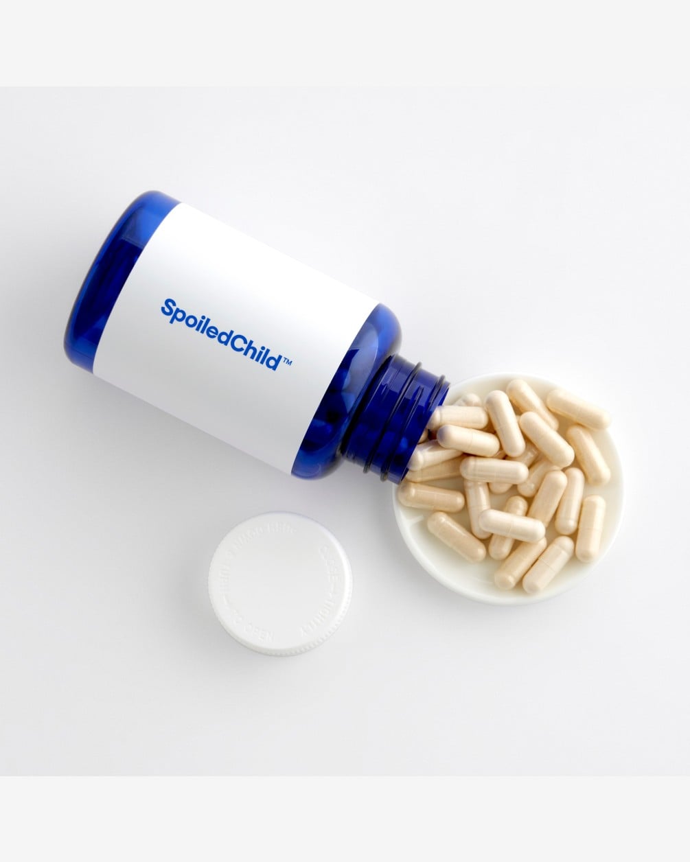 A top-down product shot of a blue SpoiledChild supplement bottle lying on its side. Several white capsules have spilled out of the bottle onto a small white circular dish. The white cap sits nearby on a clean white background.