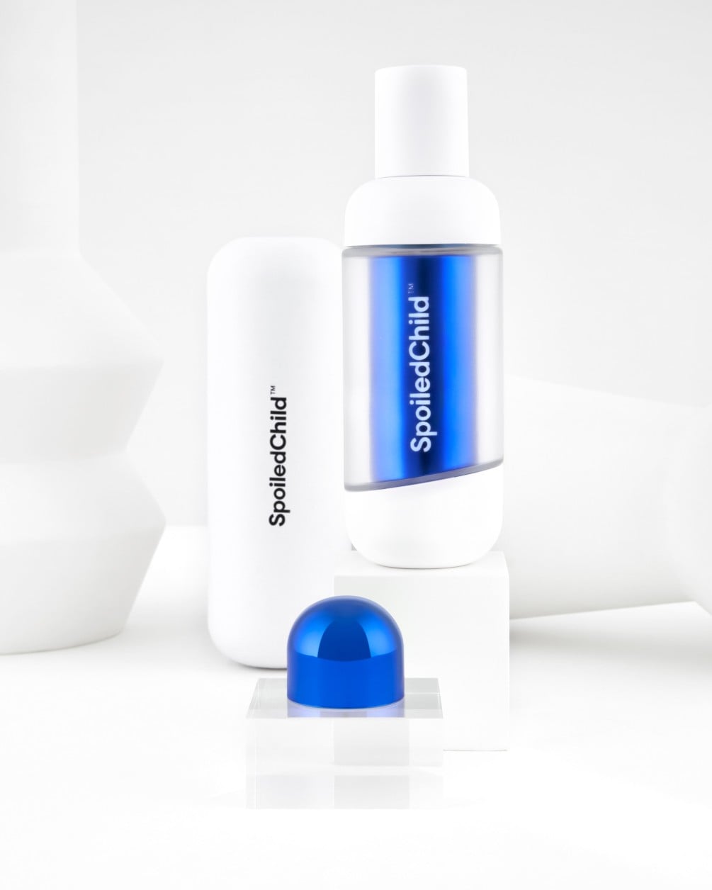 A minimalist product display on white geometric pedestals featuring SpoiledChild F38 skincare. It includes a white bottle with a clear section revealing a metallic blue label, a larger white container, and a metallic blue dome-shaped capsule.
