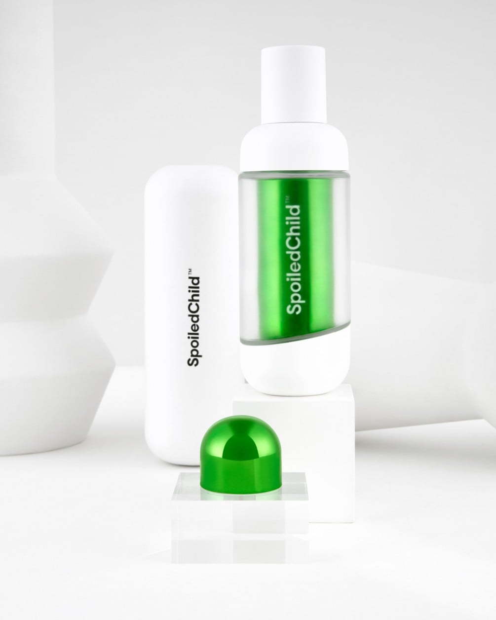 Una exhibición minimalista sobre pedestales geométricos blancos con productos SpoiledChild A35. Incluye una botella blanca con sección transparente y etiqueta azul metálico, un envase blanco más grande y una cápsula redondeada en verde metálico.