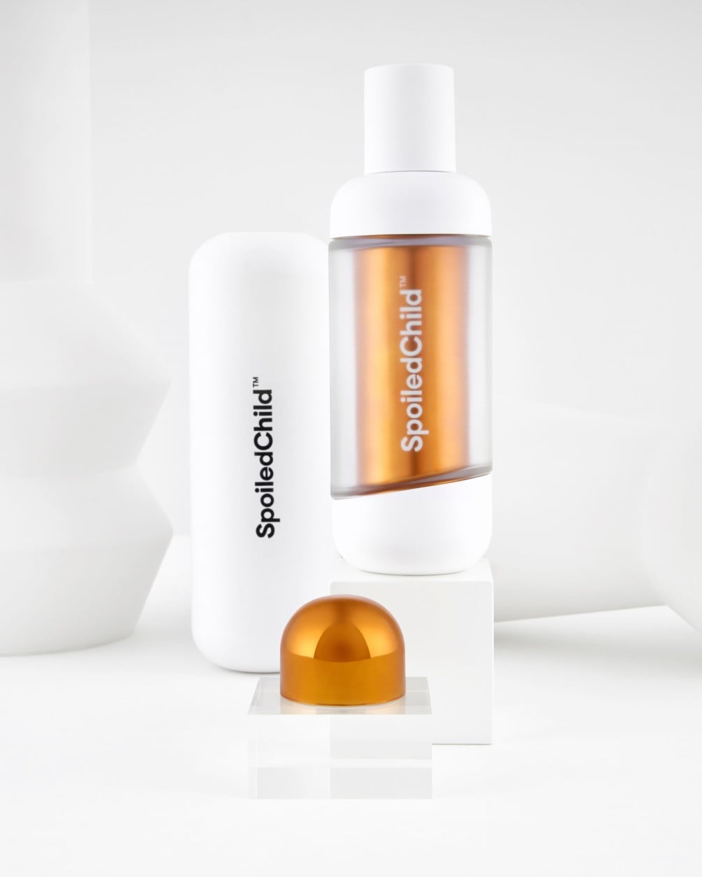 A white SpoiledChild A22 bottle with a visible copper refill capsule stands on a white block next to a solid white container and a small copper cap.
