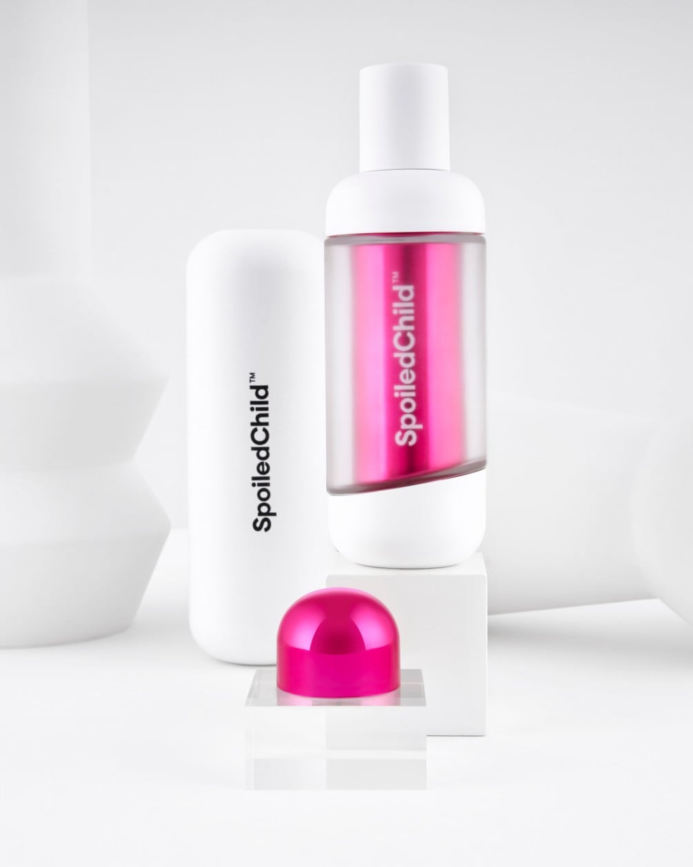 A minimalist product display on white geometric pedestals featuring SpoiledChild O36 skincare. It includes a white bottle with a clear section revealing a label, a larger white container, and a metallic pink dome-shaped capsule.