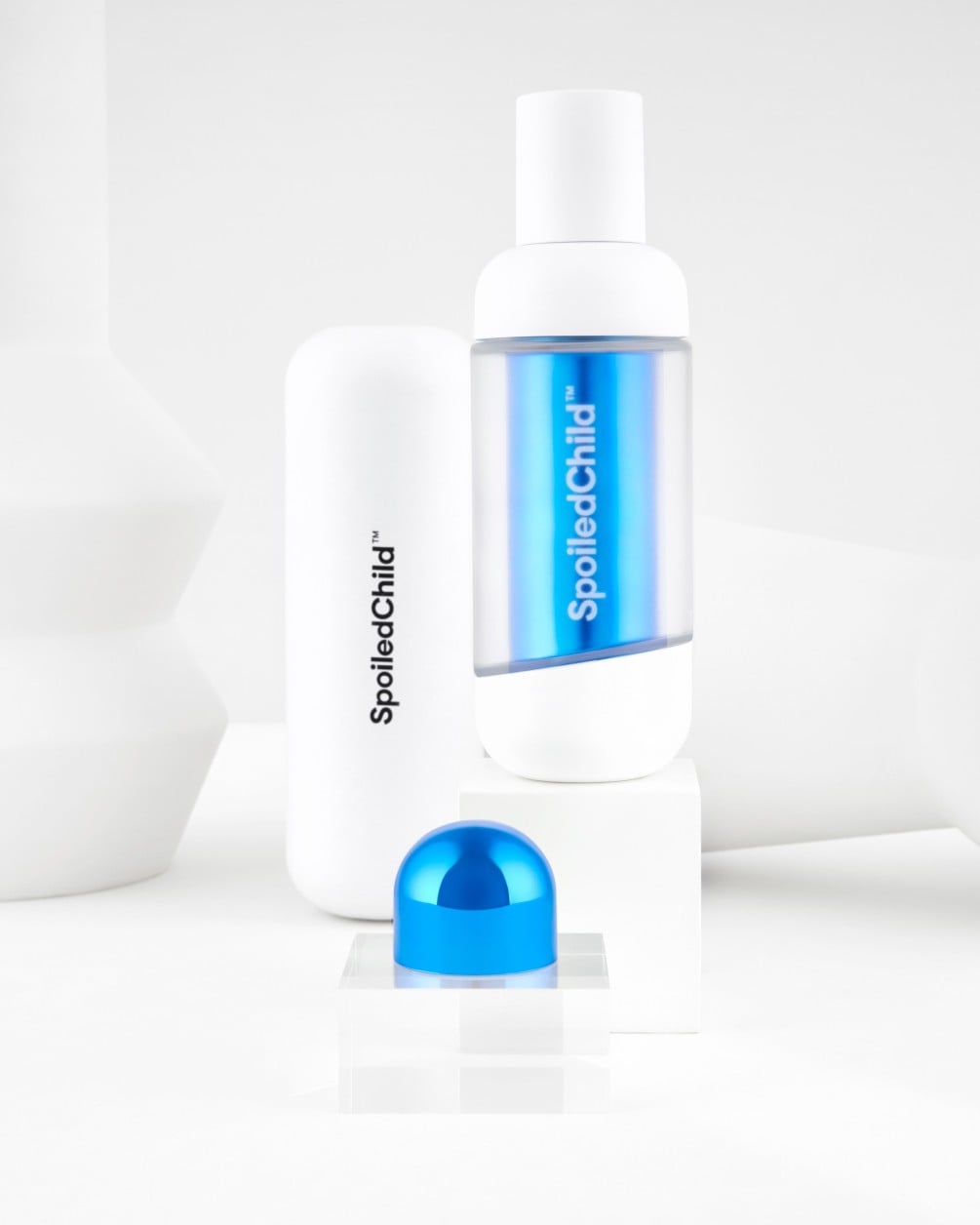 A product arrangement on white geometric pedestals featuring SpoiledChild G25 skincare items. The display includes a white bottle with a blue wrap-around label, a taller white container, and a small, metallic blue rounded capsule.