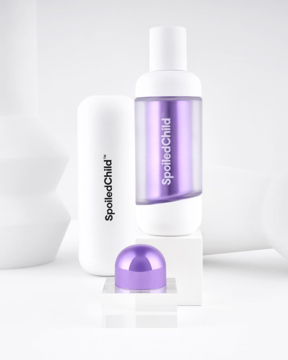 A minimalist product display on white geometric pedestals featuring SpoiledChild C29 skincare. It includes a white bottle with a clear section revealing a label, a larger white container, and a metallic violet dome-shaped capsule.