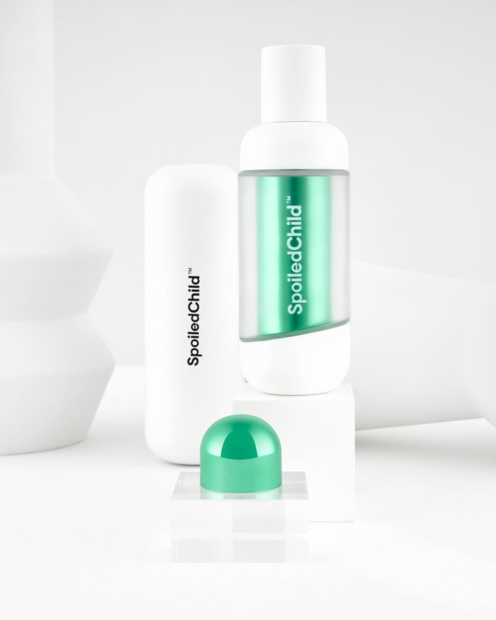 A minimalist product display on white geometric pedestals featuring SpoiledChild S34 skincare. It includes a white bottle with a clear section revealing a metallic blue label, a larger white container, and a metallic green dome-shaped capsule.