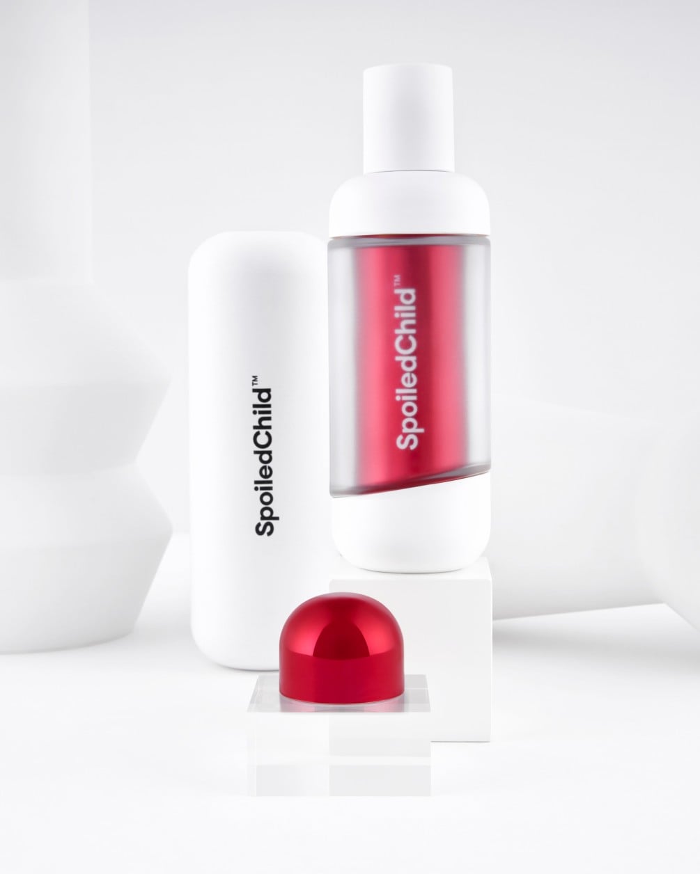 A minimalist product display on white geometric pedestals featuring SpoiledChild D35+ skincare. It includes a white bottle with a clear section revealing a label, a larger white container, and a metallic red dome-shaped capsule.