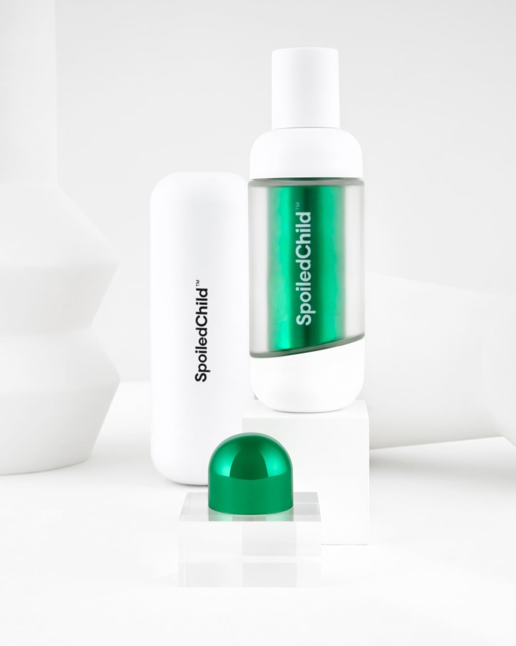 A product icon featuring a white SpoiledChild M27 bottle with a clear body revealing a metallic teal refill capsule inside. In the background, a second white container stands tall, and a small metallic teal cap rests in the foreground on a white pedestal.