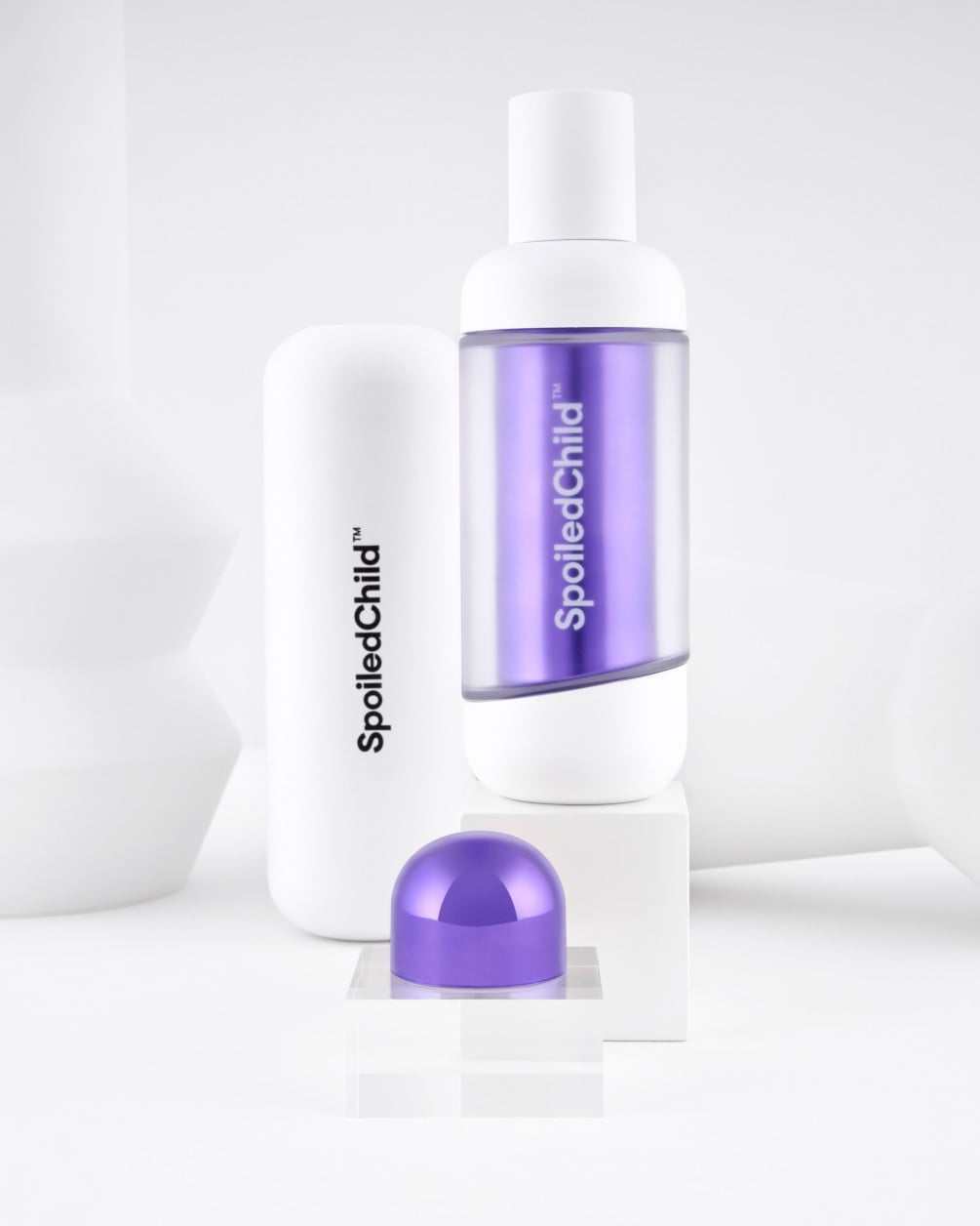 A minimalist product display on white geometric pedestals featuring SpoiledChild L28 skincare. It includes a white bottle with a clear section revealing a label, a larger white container, and a metallic dark violet dome-shaped capsule.