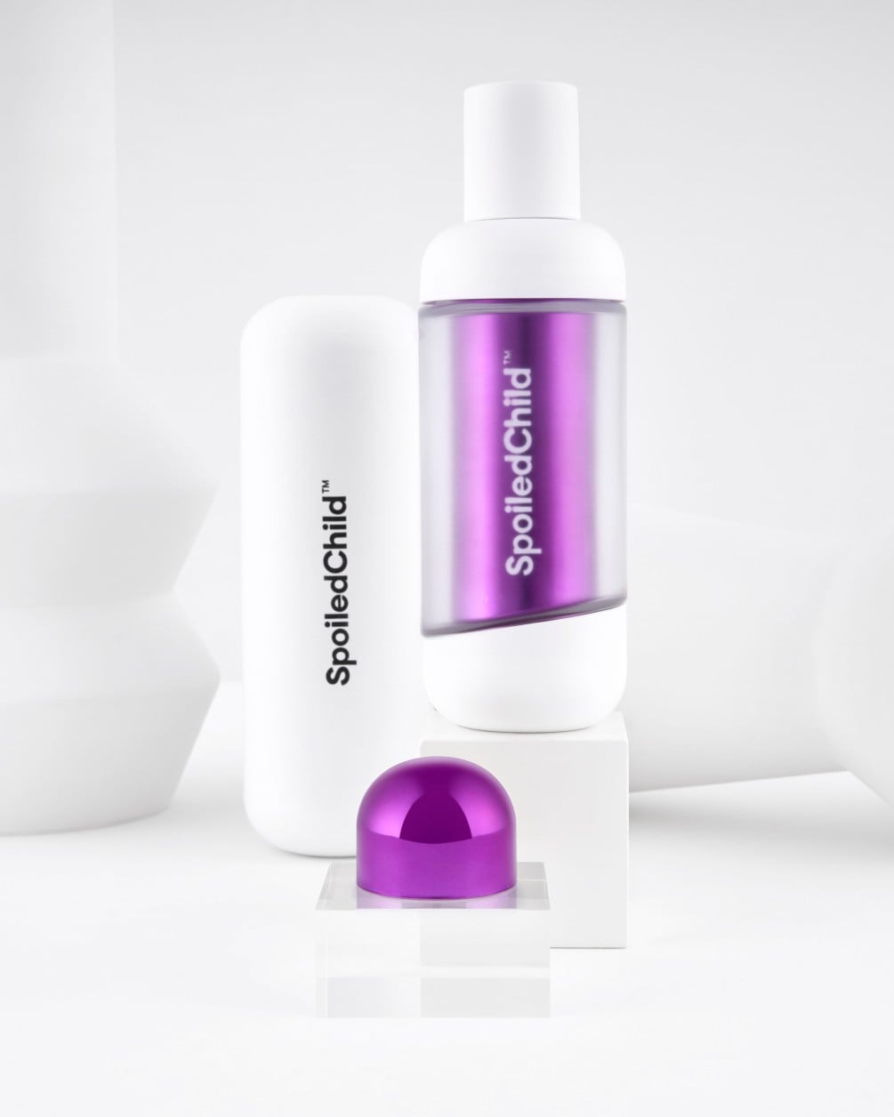 A minimalist product display on white geometric pedestals featuring SpoiledChild S27+ skincare. It includes a white bottle with a clear section revealing a label, a larger white container, and a metallic purple dome-shaped capsule.