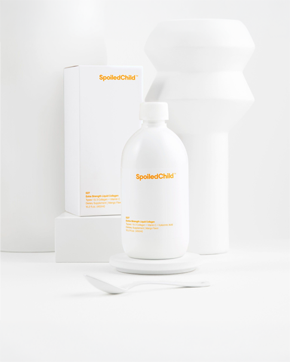 A minimalist lifestyle shot featuring the white SpoiledChild mango liquid collagen bottle and its product box arranged against a clean, white geometric background.