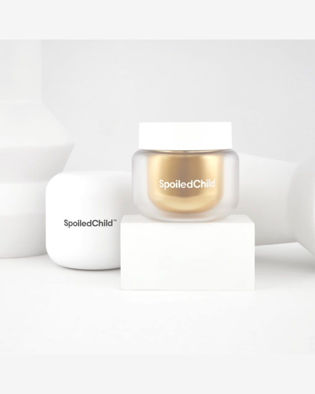 A SpoiledChild S24 product jar with a gold inner container and white cap sits on a white block. Its white product box and a white sculptural vase are visible in the background against a clean white setting.