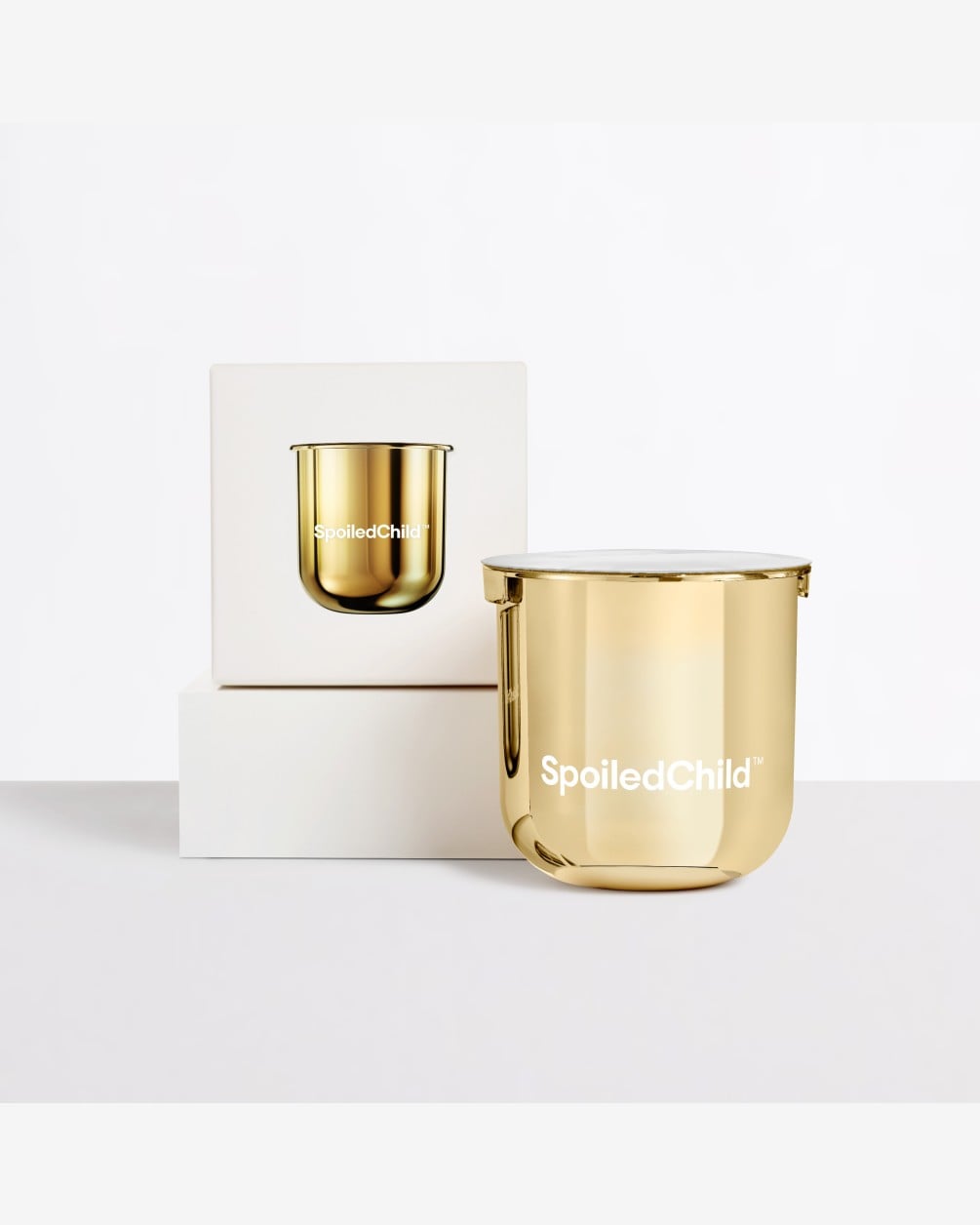 A reflective gold SpoiledChild S24 refill capsule stands on a white surface next to its product box. The box, which features an image of the capsule, is elevated on a white block against a clean white background.