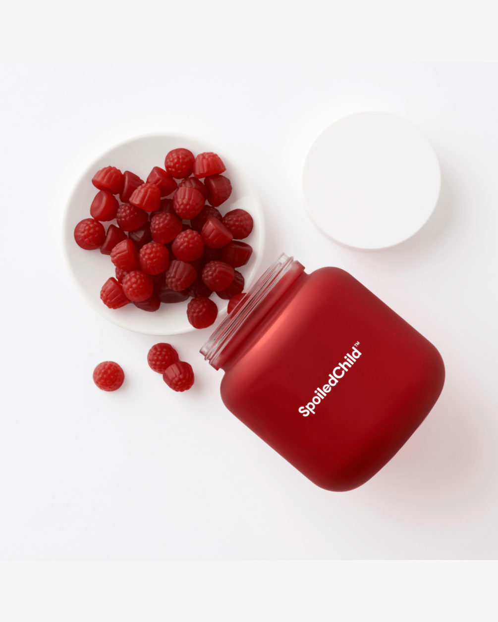 A top-down view of a matte red SpoiledChild L30 jar lying on its side next to a small white dish filled with red, raspberry-shaped gummy supplements.