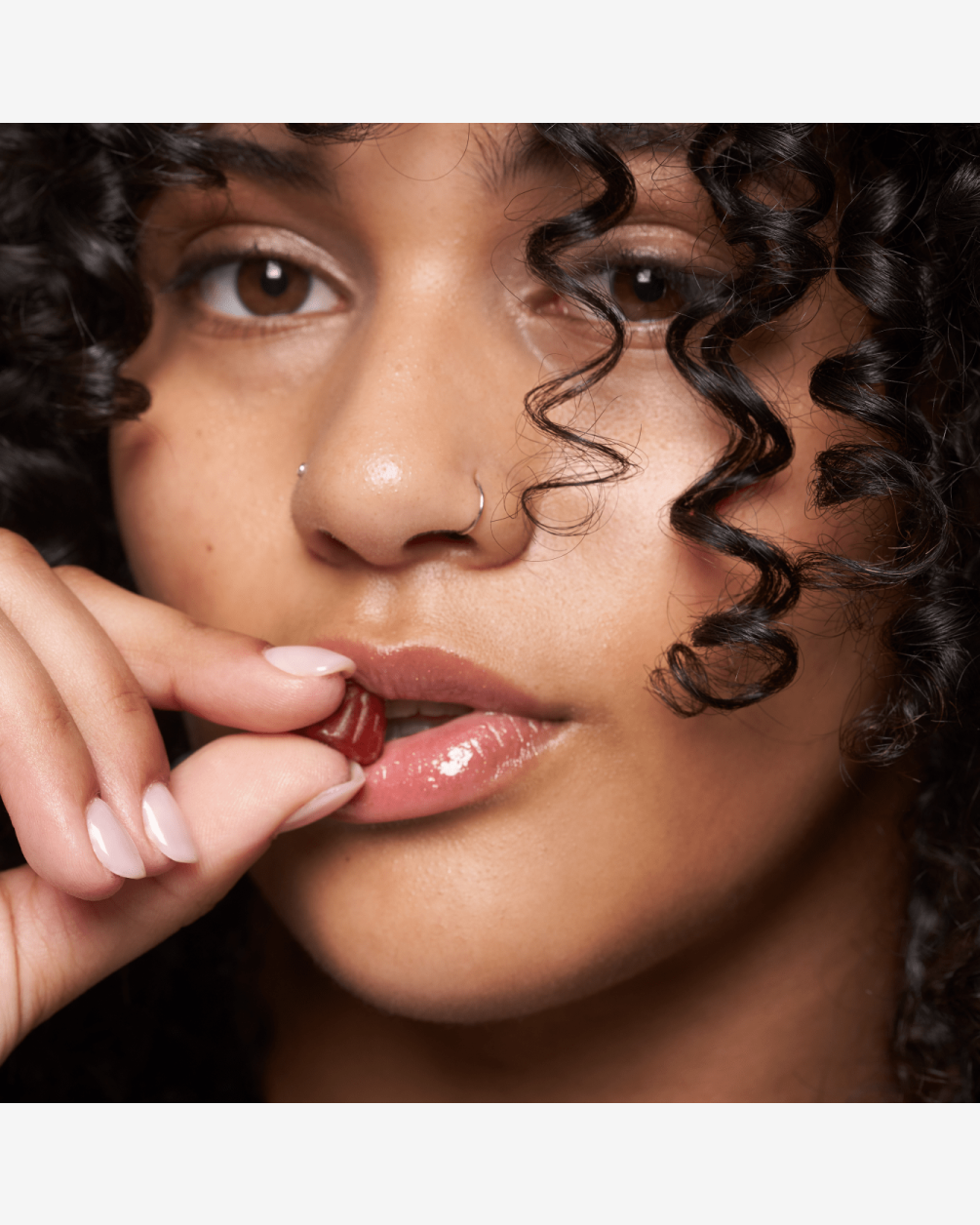 A close-up portrait of a woman with dark curly hair and a nose ring. She is holding a single red gummy L30 supplement to her lips, which have a glossy finish.