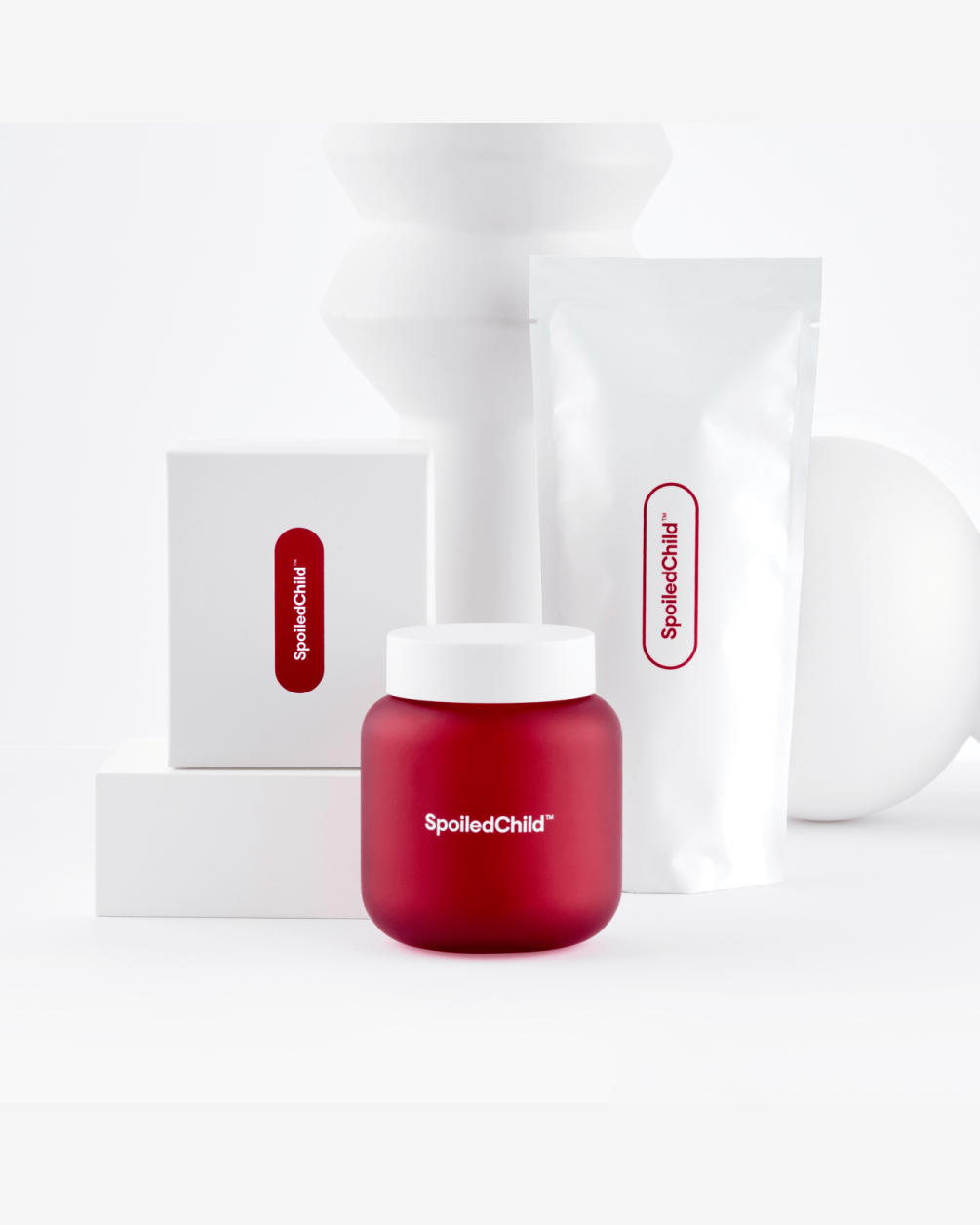 A minimalist product display featuring a matte red SpoiledChild L30 jar in the foreground. In the background are a white box and a white refill pouch, both featuring red branding, arranged on white geometric blocks.