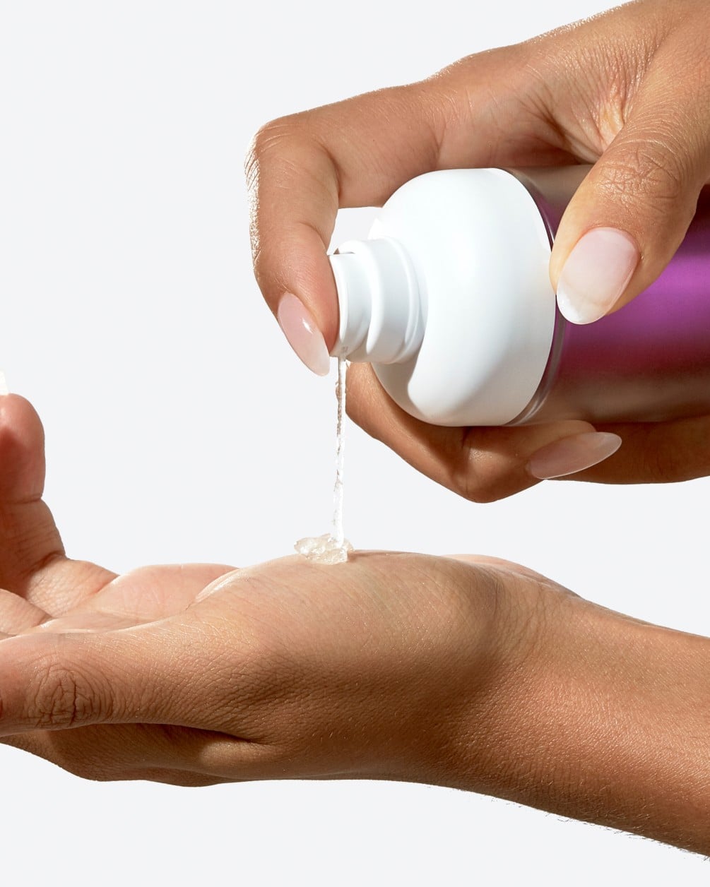 A close-up action shot showing a person with a deeper skin tone dispensing a creamy liquid from a white SpoiledChild S27+ pump bottle. A small amount of the product is shown landing in the palm of their other hand.