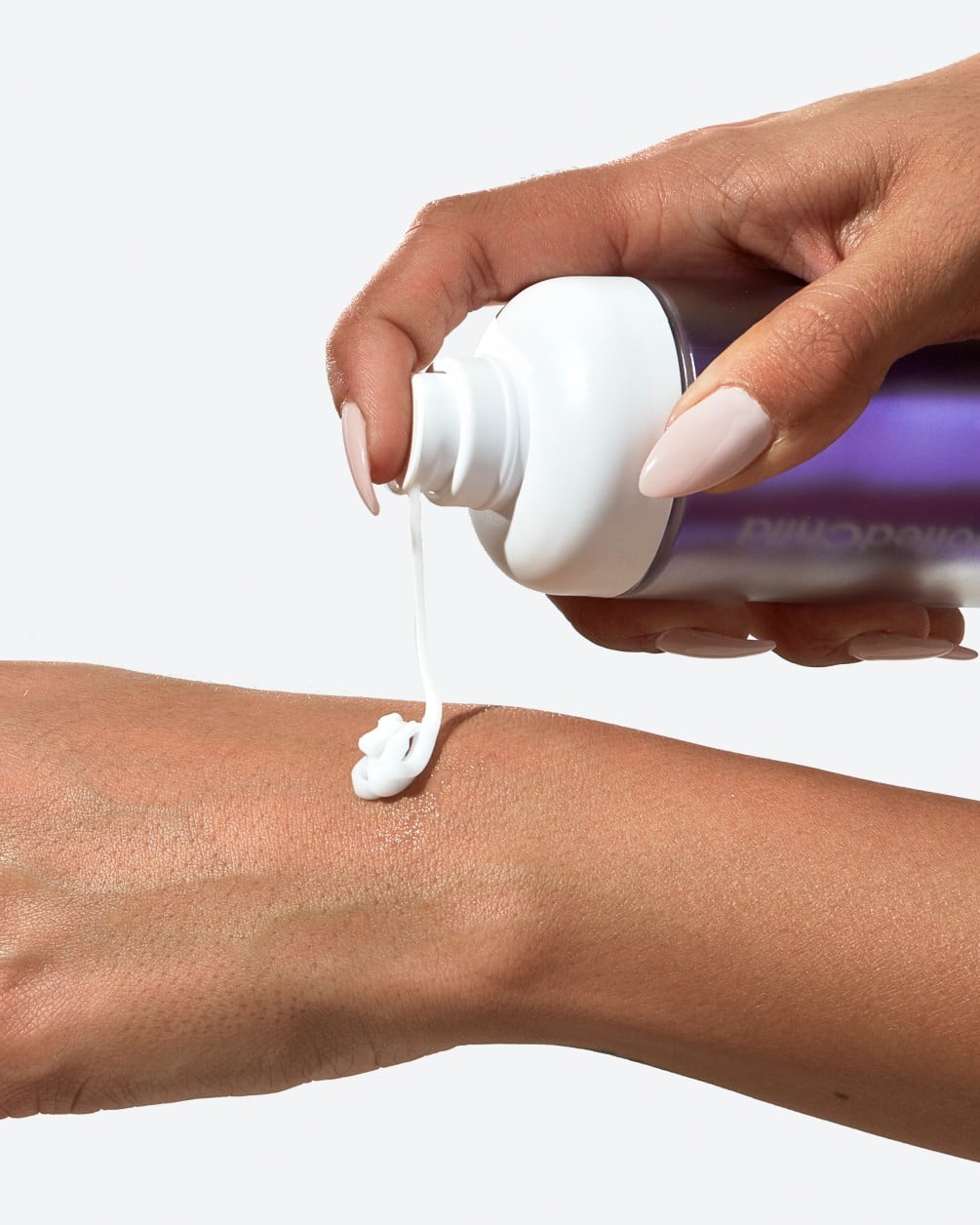 A close-up action shot showing a person with a deeper skin tone dispensing a creamy liquid from a white SpoiledChild L28 pump bottle. A small amount of the product is shown landing in the palm of their other hand.