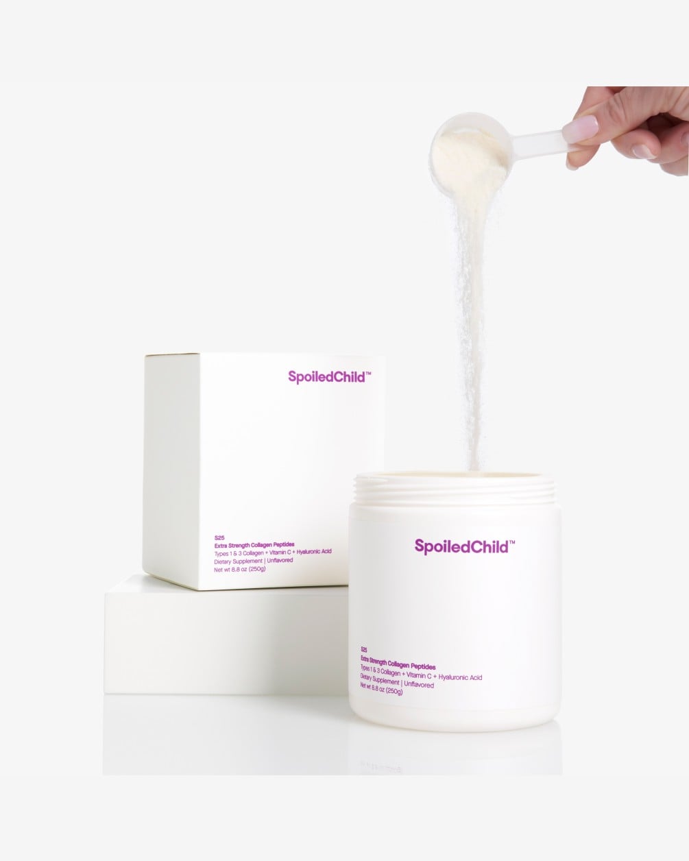 A person's hand uses a white scoop to pour a stream of SpoiledChild S25 Extra Strength Collagen Peptides powder into its large white tub. The product's matching white box is elevated on a block in the background against a clean white setting.