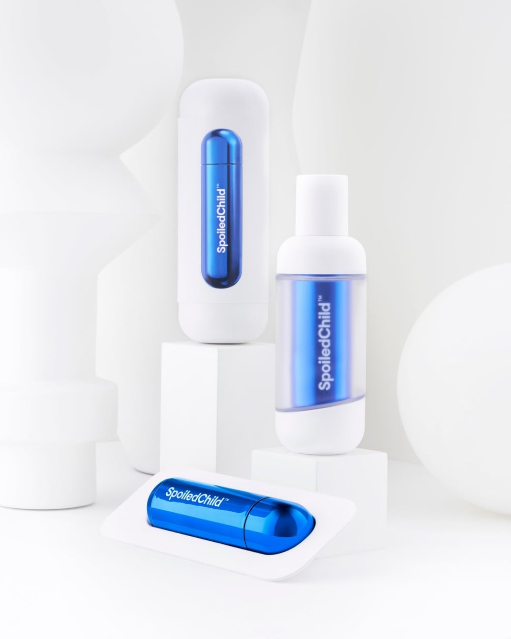A minimalist product arrangement of SpoiledChild F38 skincare items on white geometric pedestals against a clean white background. The set includes two white bottles and a separate metallic blue refill.