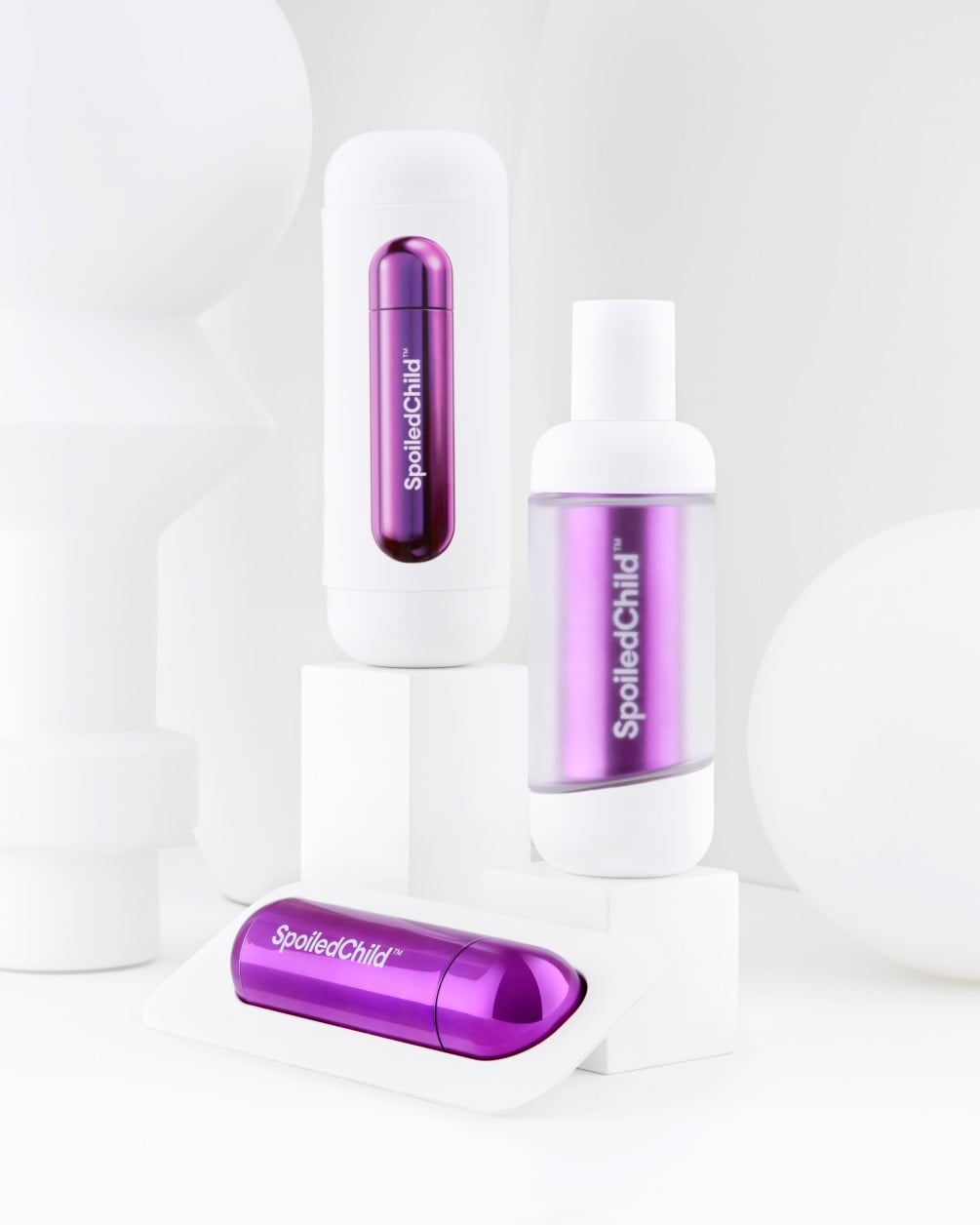 A minimalist product arrangement of SpoiledChild S27+ skincare items on white geometric pedestals against a clean white background. The set includes two white bottles and a separate metallic purple refill.