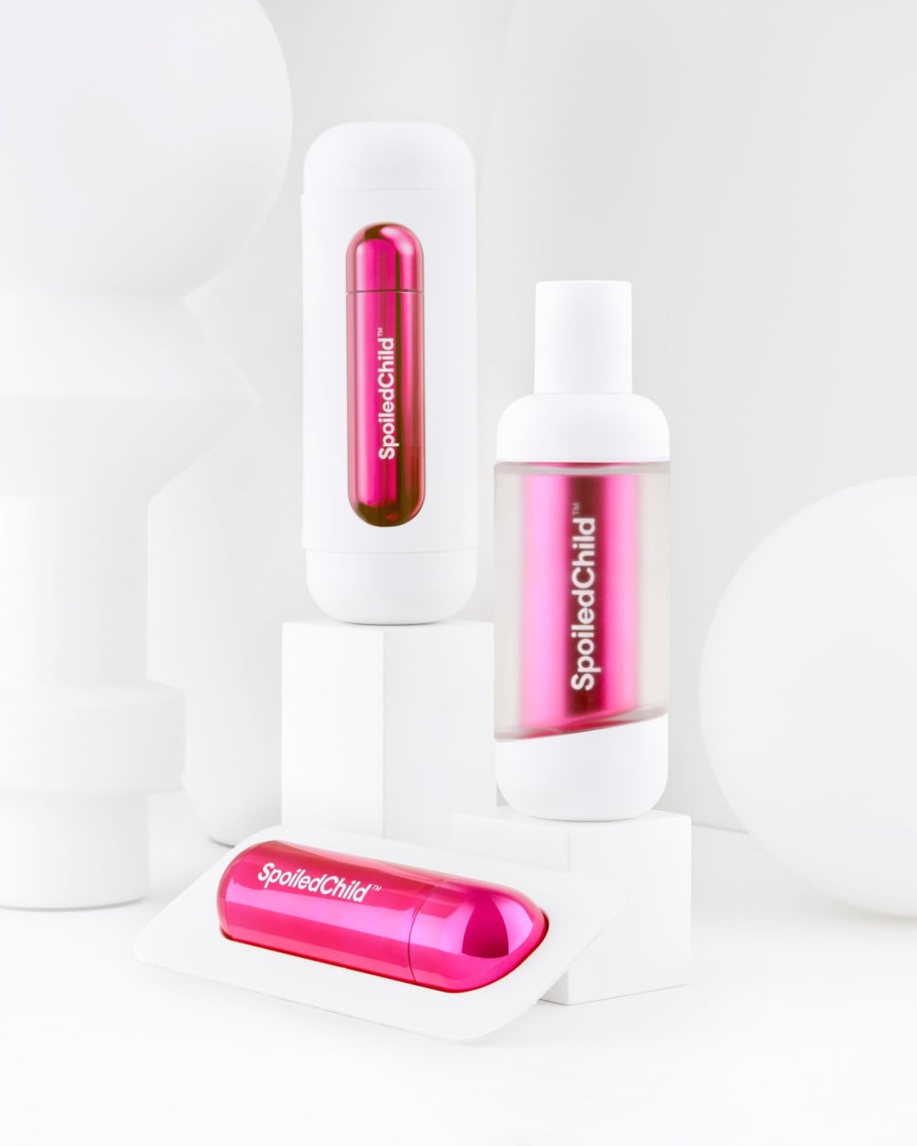 A minimalist product arrangement of SpoiledChild O36 skincare items on white geometric pedestals against a clean white background. The set includes two white bottles and a separate metallic pink refill.