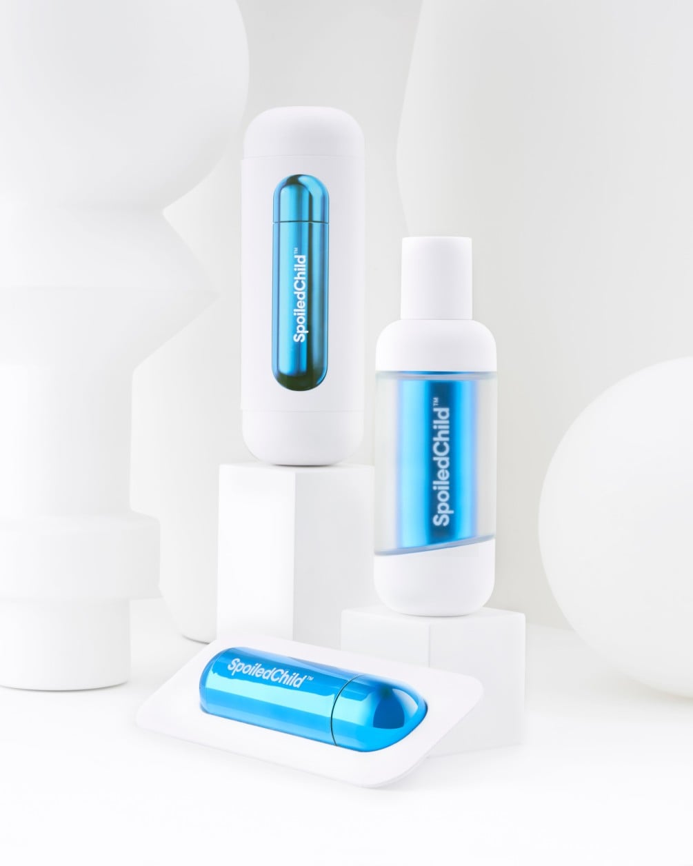 A product arrangement on white pedestals featuring SpoiledChild G25 items. The display includes a white container and a bottle, both showing metallic blue wrap-around labels through clear sections. A separate metallic blue refil lies horizontally.