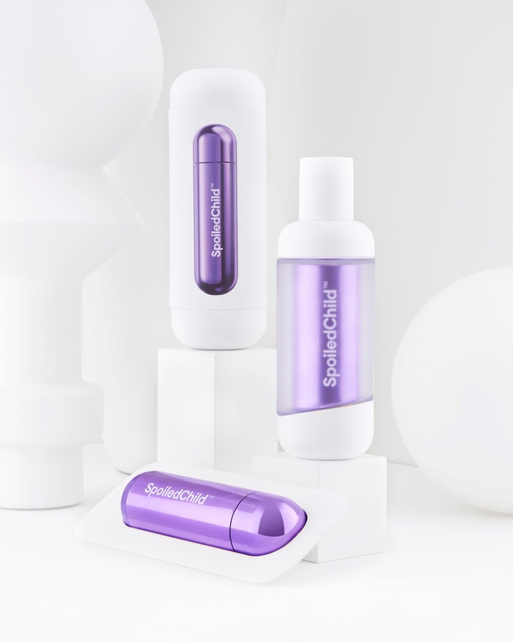 A minimalist product arrangement of SpoiledChild C29 skincare items on white geometric pedestals against a clean white background. The set includes two white bottles and a separate metallic violet refill.
