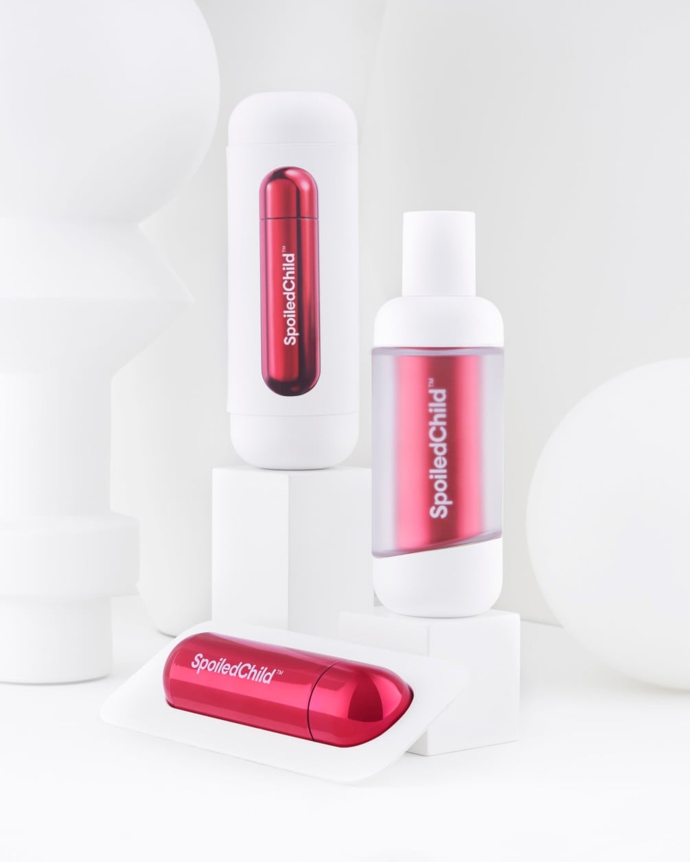 A minimalist product arrangement of SpoiledChild D35+ skincare items on white geometric pedestals against a clean white background. The set includes two white bottles and a separate metallic red refill.