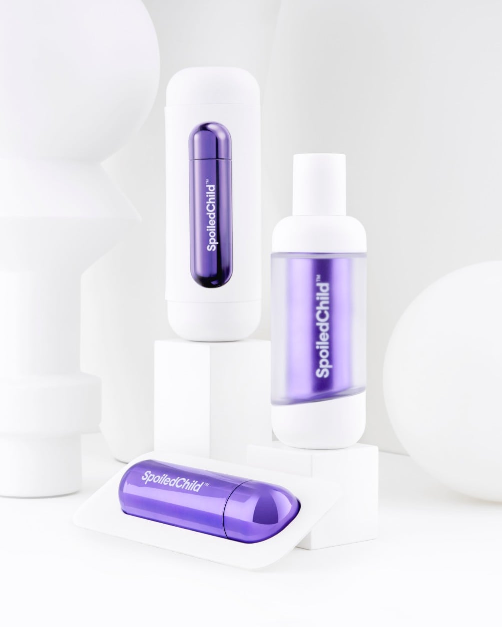 A minimalist product arrangement of SpoiledChild L28 skincare items on white geometric pedestals against a clean white background. The set includes two white bottles and a separate metallic dark violet refill.