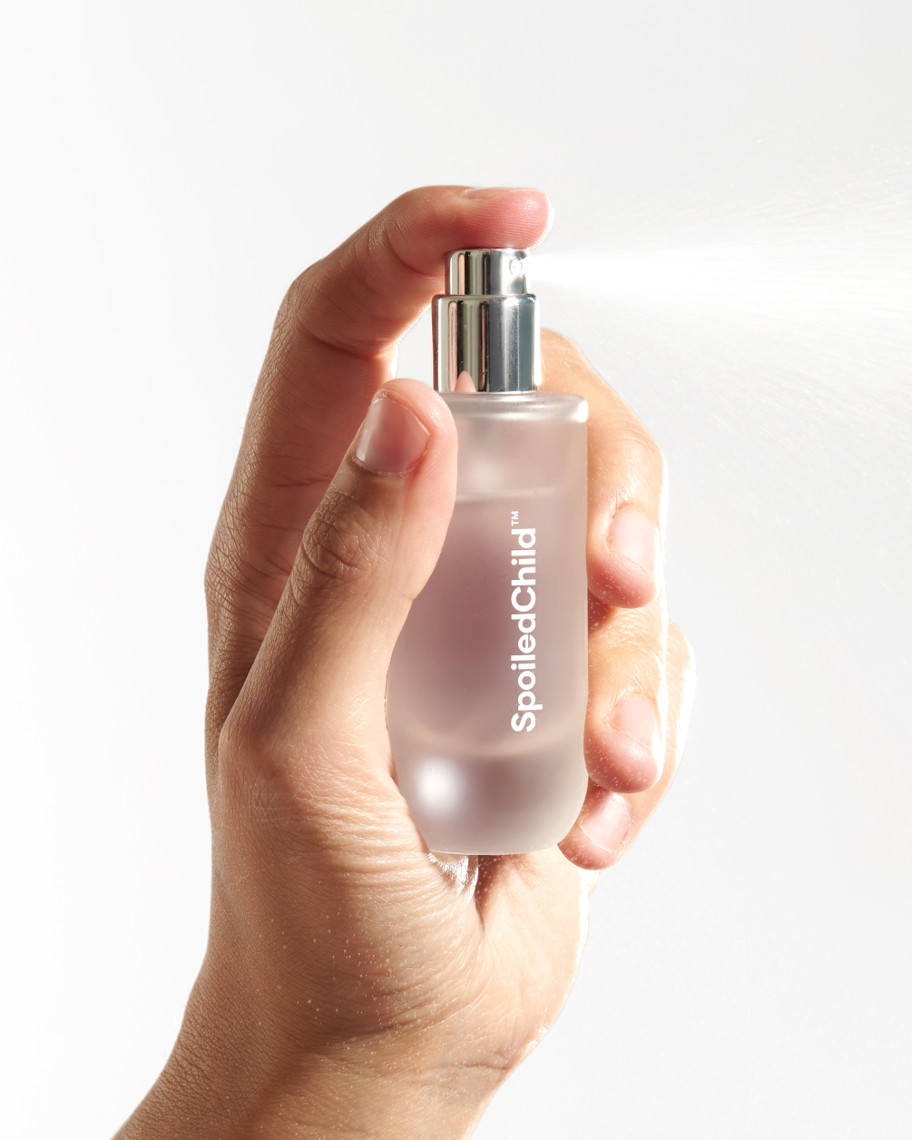 A hand holding a frosted glass spray bottle, dispensing a fine mist of liquid against a white background.