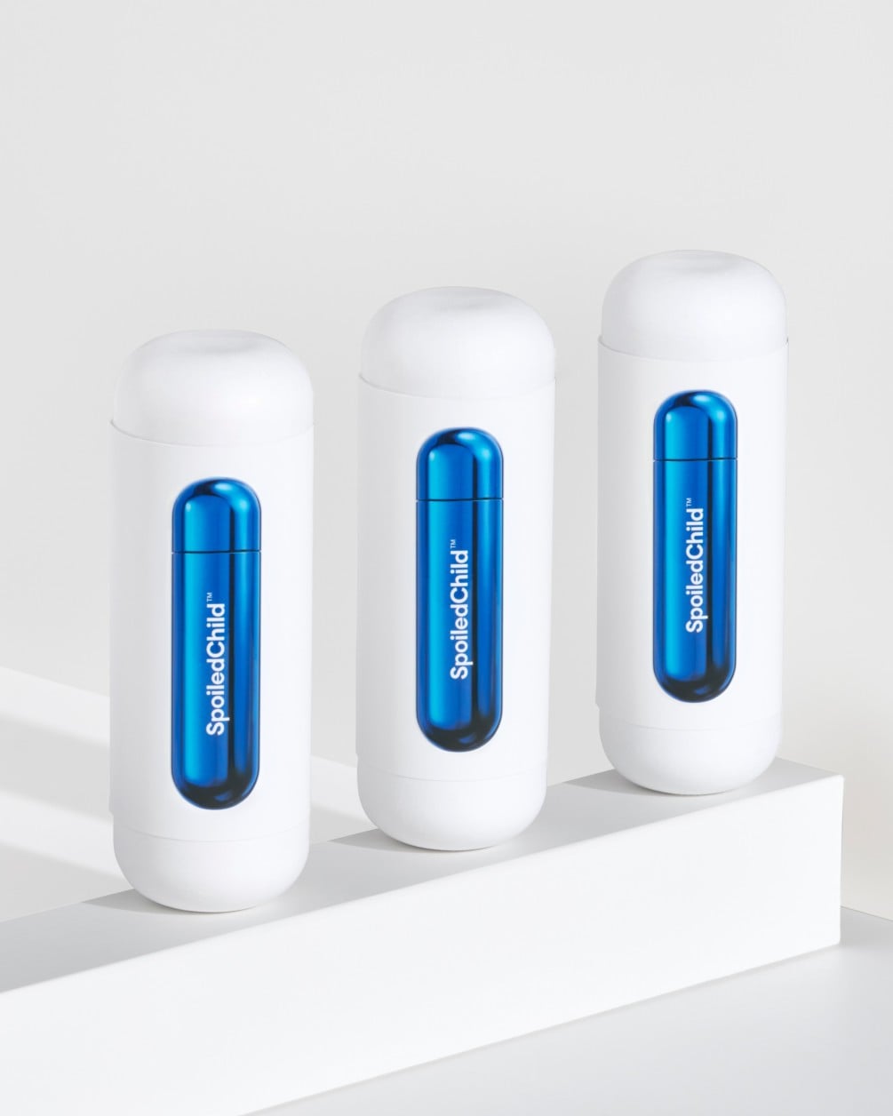 Three white SpoiledChild F38 skincare bottles arranged in a diagonal row on a white minimalist ledge. Each bottle features a clear capsule-shaped window in the center that reveals a vibrant, metallic blue internal refill.