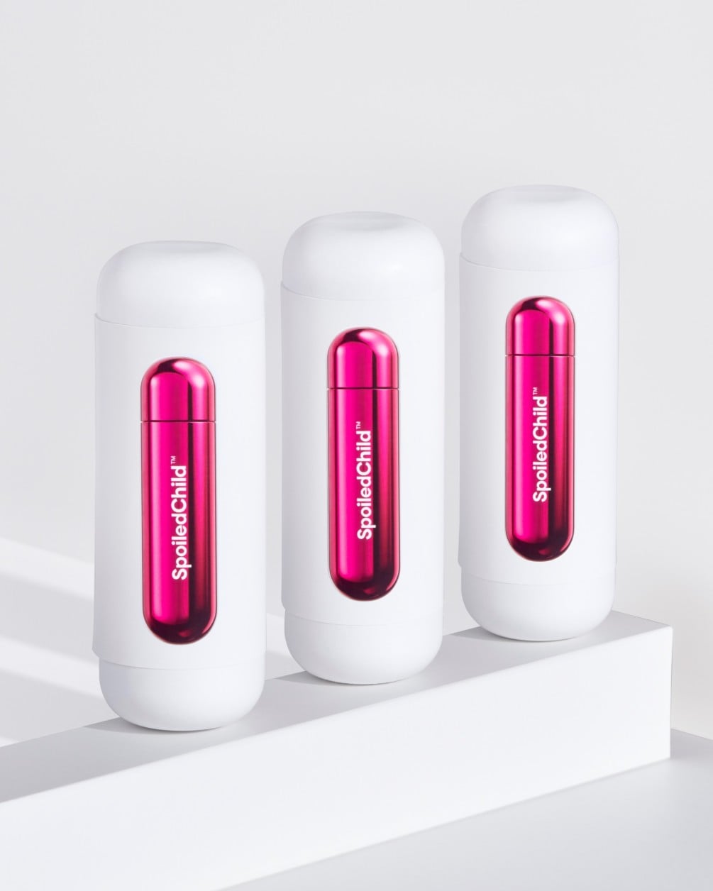 Three white SpoiledChild O36 skincare bottles arranged in a diagonal row on a white minimalist ledge. Each bottle features a clear capsule-shaped window in the center that reveals a vibrant, metallic pink internal refill.