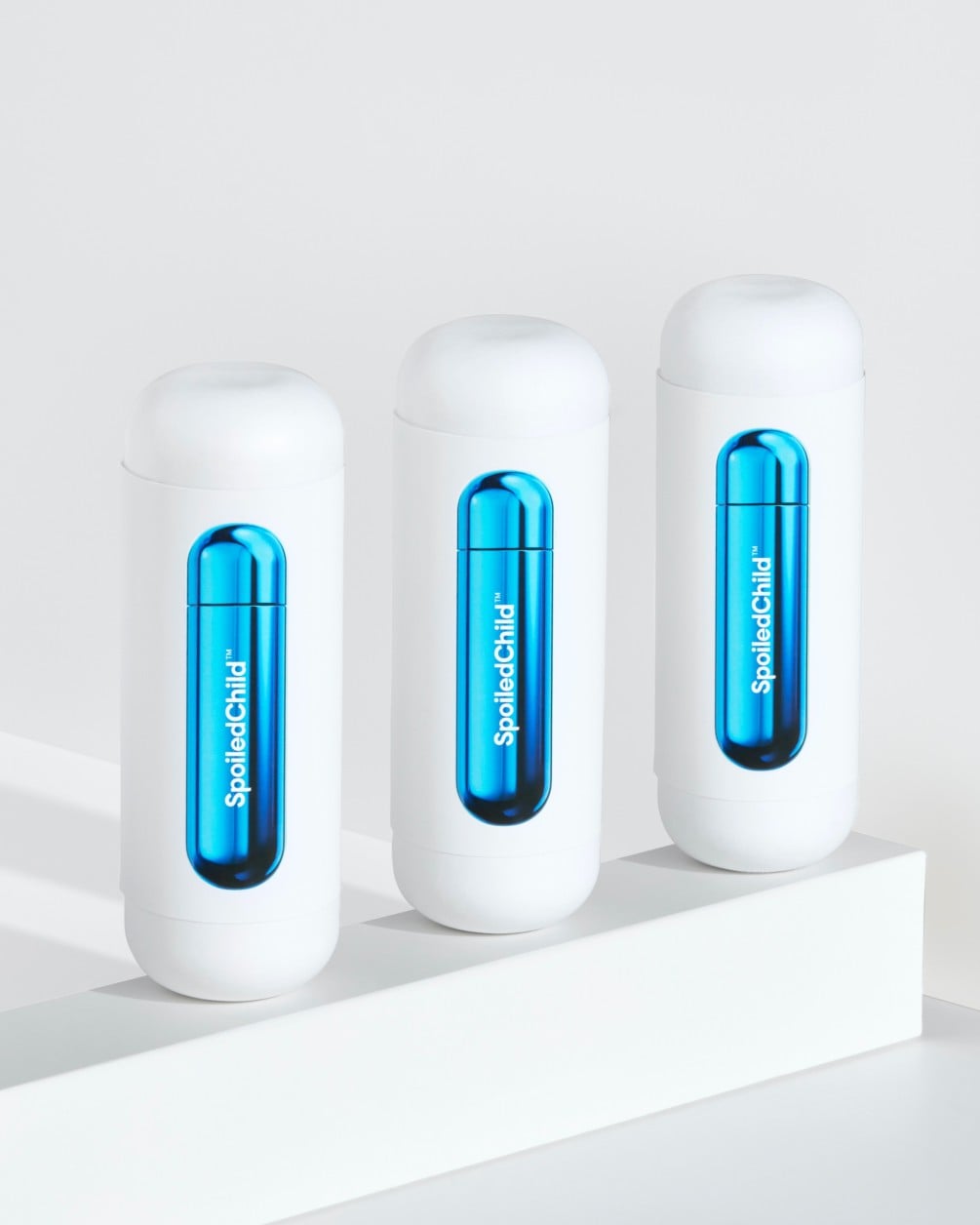 A minimalist product arrangement featuring three white SpoiledChild G25 containers lined up on a white ledge. Each container features a sleek, metallic blue wrap-around label with the brand name printed vertically.