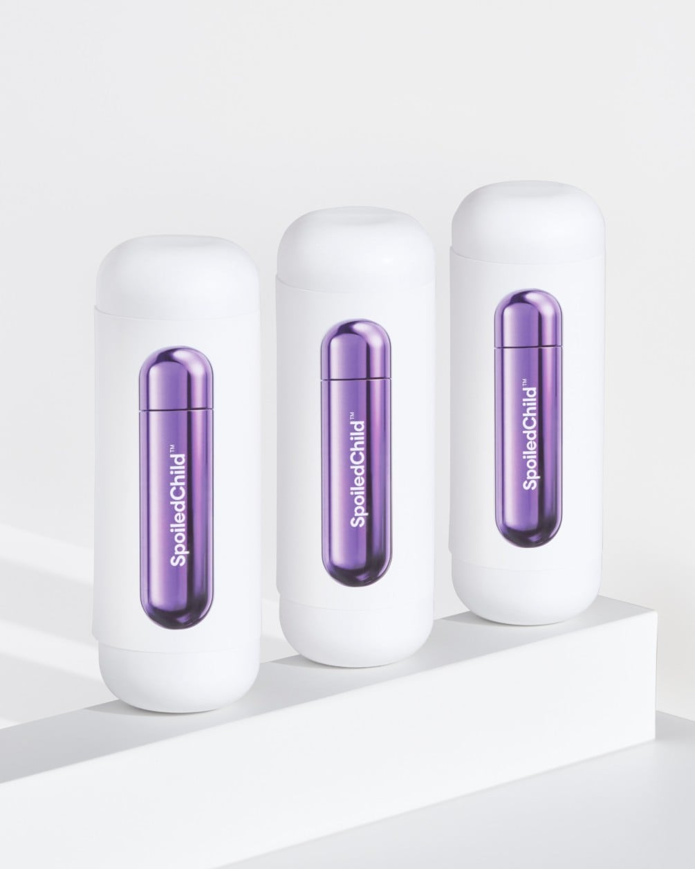 Three white SpoiledChild C29 skincare bottles arranged in a diagonal row on a white minimalist ledge. Each bottle features a clear capsule-shaped window in the center that reveals a vibrant, metallic violet internal refill.