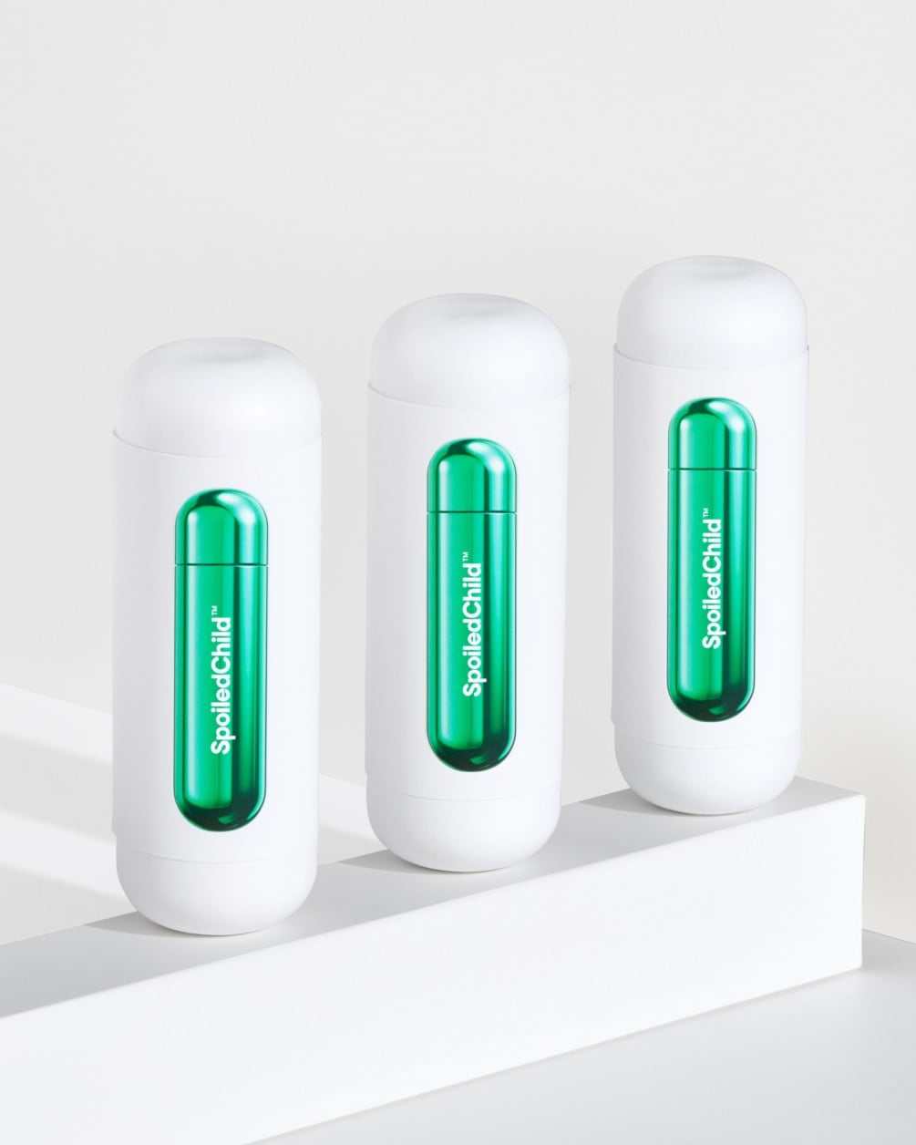Three white SpoiledChild S34 skincare bottles arranged in a diagonal row on a white minimalist ledge. Each bottle features a clear capsule-shaped window in the center that reveals a vibrant, metallic green internal refill.