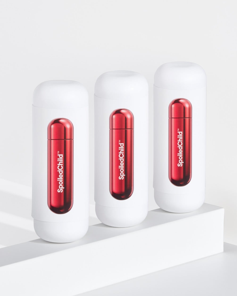 Three white SpoiledChild D35+ skincare bottles arranged in a diagonal row on a white minimalist ledge. Each bottle features a clear capsule-shaped window in the center that reveals a vibrant, metallic red internal refill.