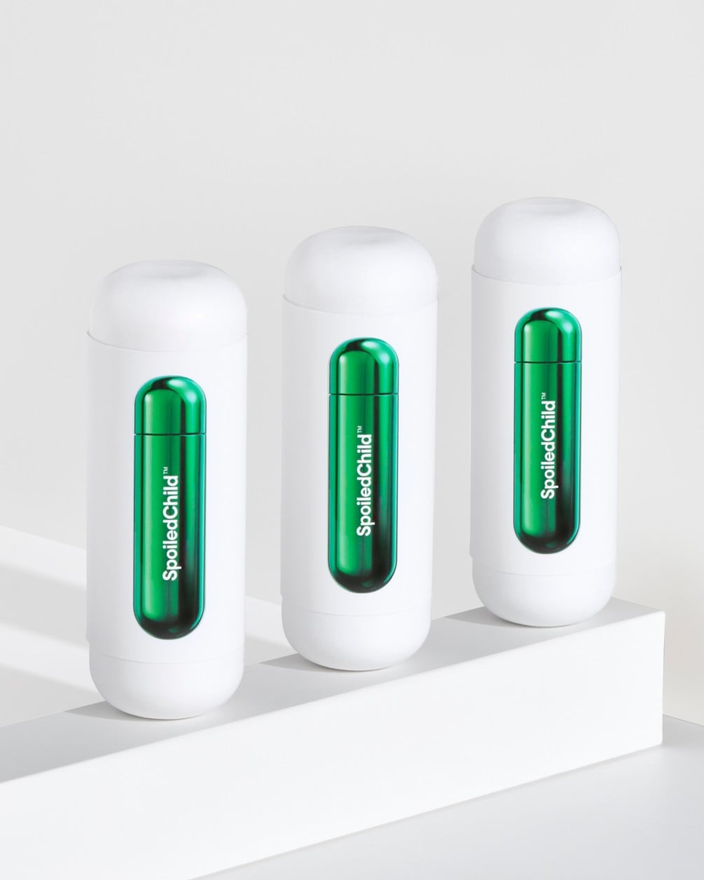 A minimalist product arrangement featuring three white SpoiledChild M27 containers lined up on a white ledge. Each container features a sleek, metallic green wrap-around label with the brand name printed vertically.