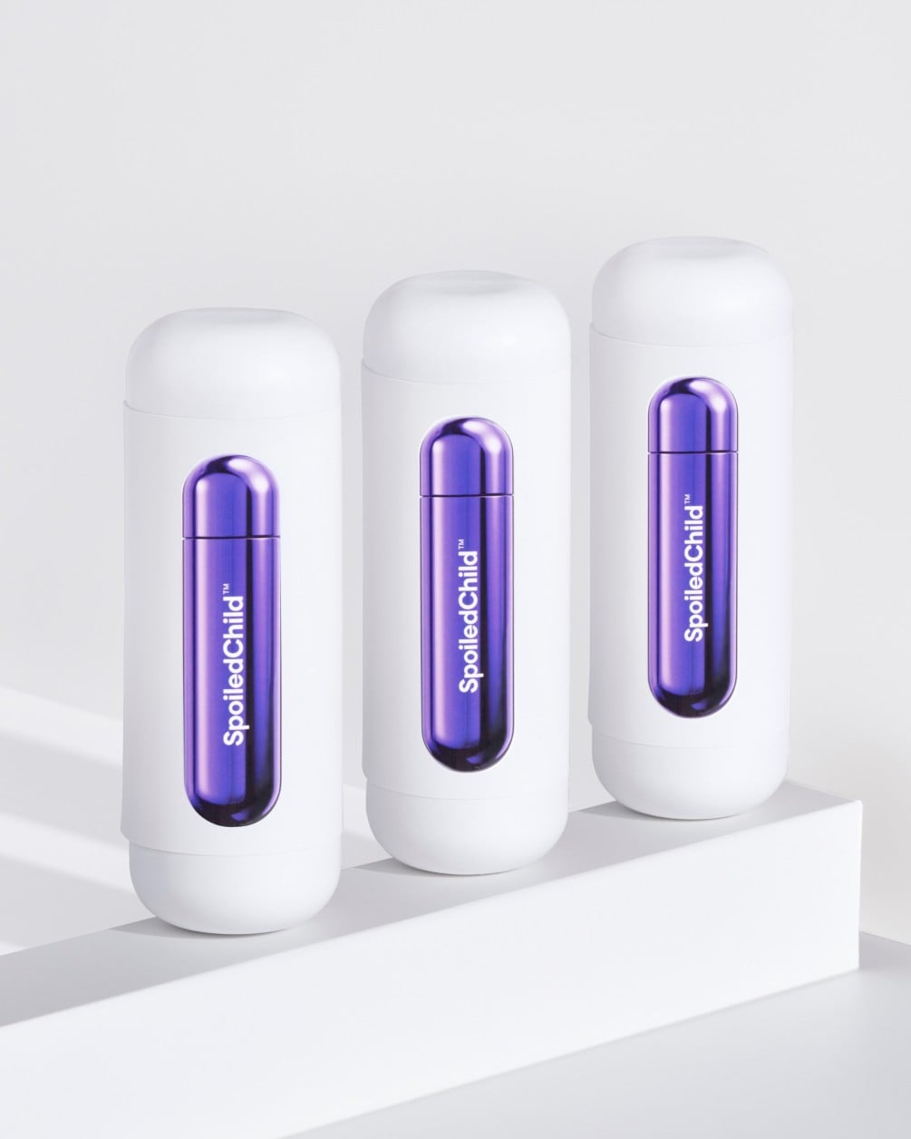 Three white SpoiledChild L28 skincare bottles arranged in a diagonal row on a white minimalist ledge. Each bottle features a clear capsule-shaped window in the center that reveals a vibrant, metallic dark violet internal refill.