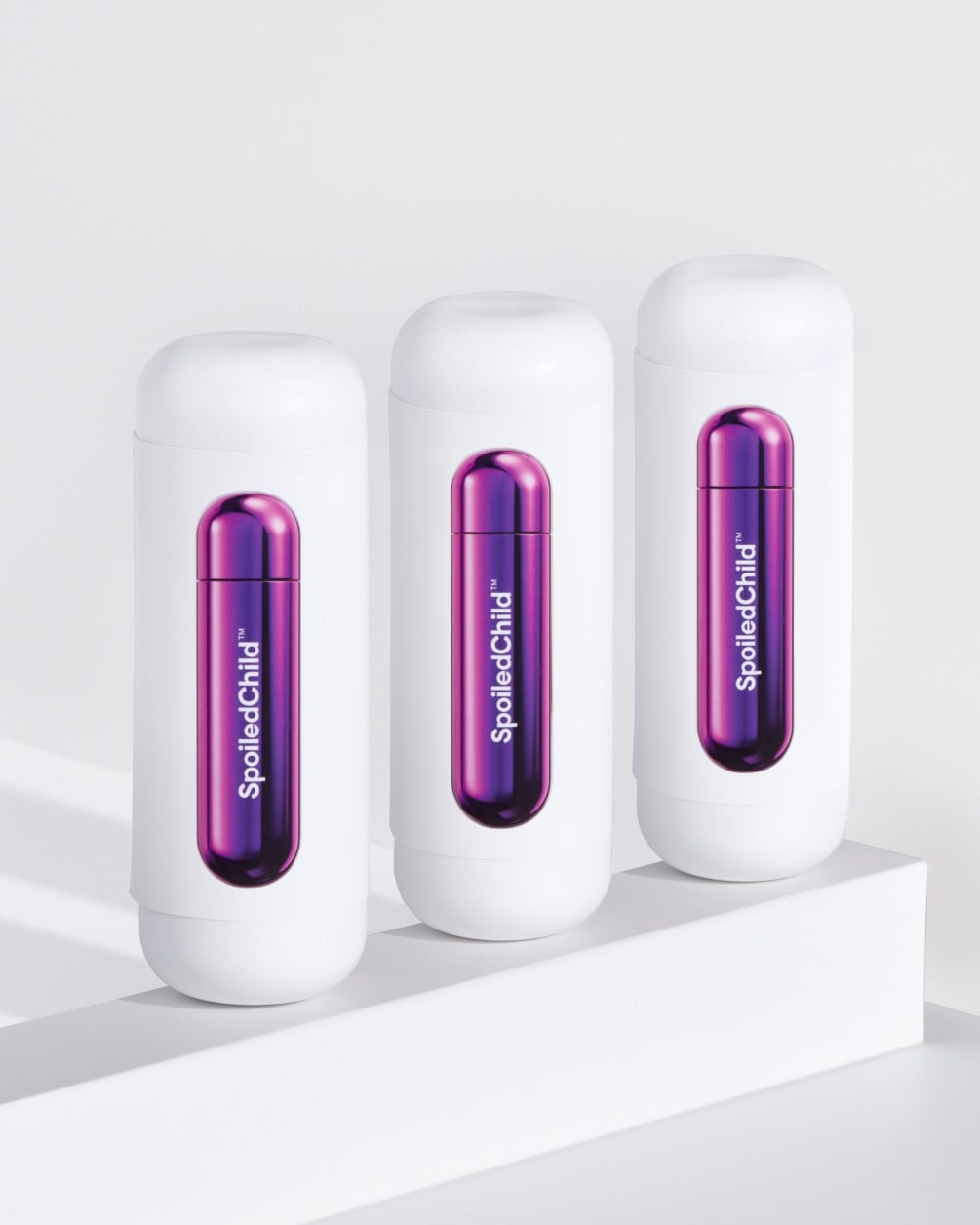Three white SpoiledChild S27+ skincare bottles arranged in a diagonal row on a white minimalist ledge. Each bottle features a clear capsule-shaped window in the center that reveals a vibrant, metallic purple internal refill.