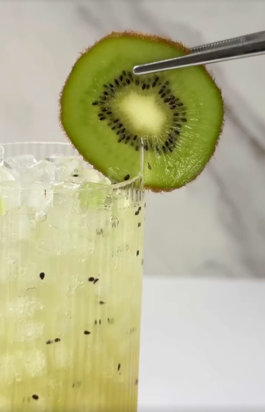 Glass of pale green kiwi gin cocktail over crushed ice, garnished with a fresh kiwi slice