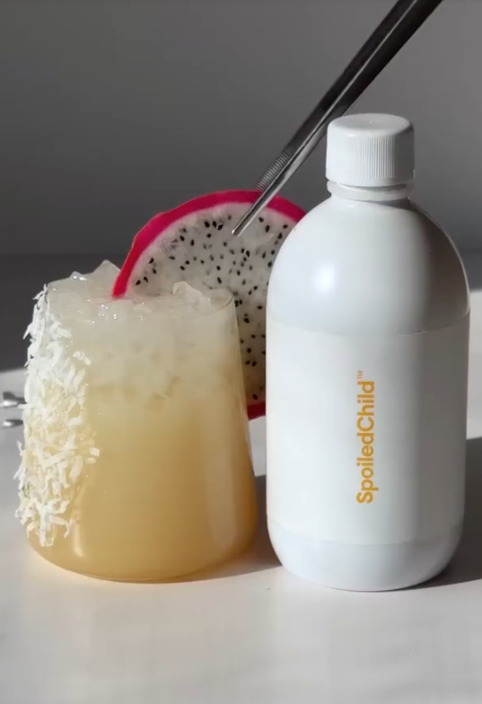 Creamy lychee collagen margarita mocktail with a coconut-flake rim and dragon-fruit slice, next to a SpoiledChild E27 bottle
